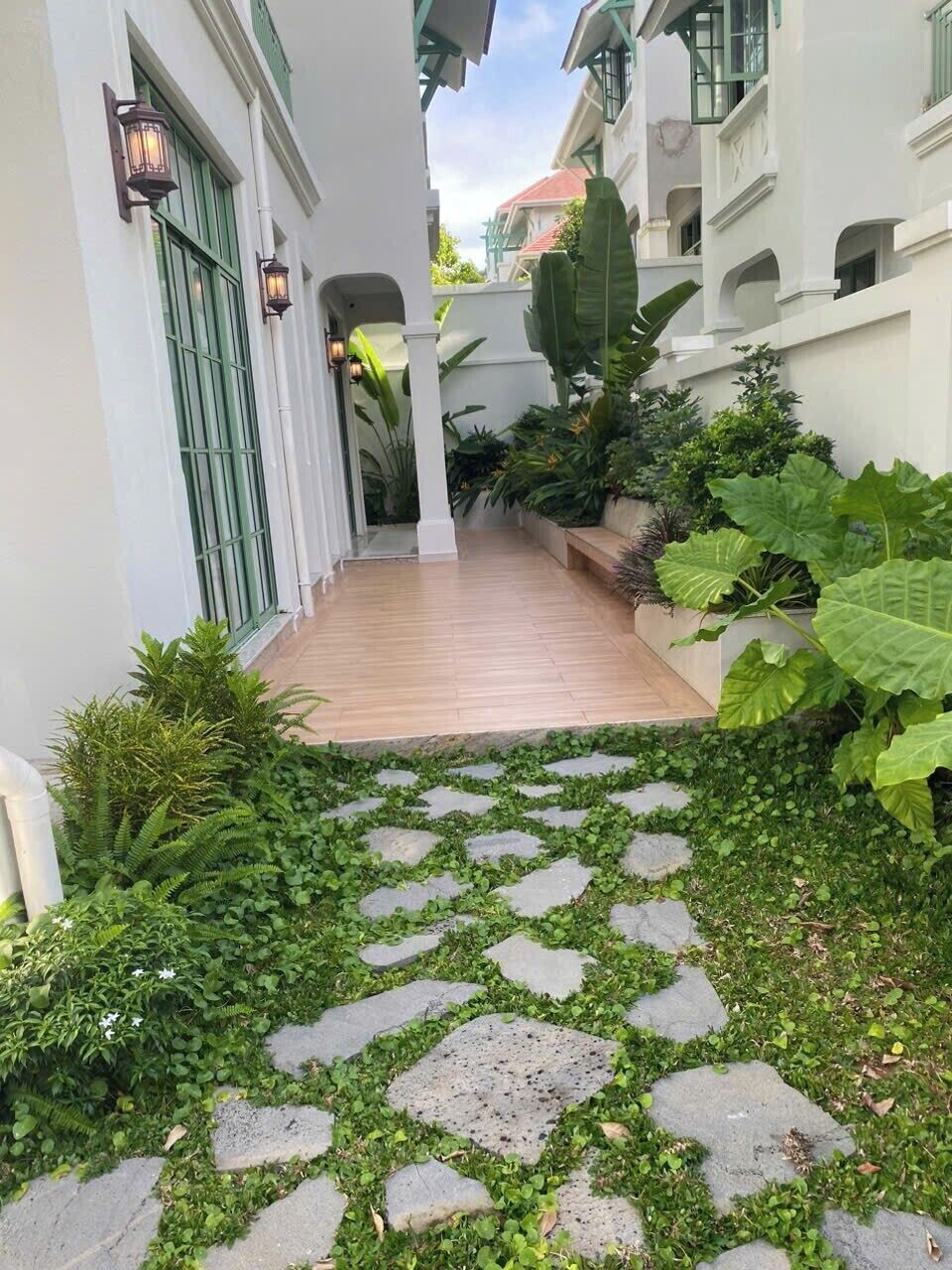 Un sendero estrecho con baldosas de piedra y abundante vegetación, bordeado por edificios blancos con detalles verdes y faroles tradicionales.