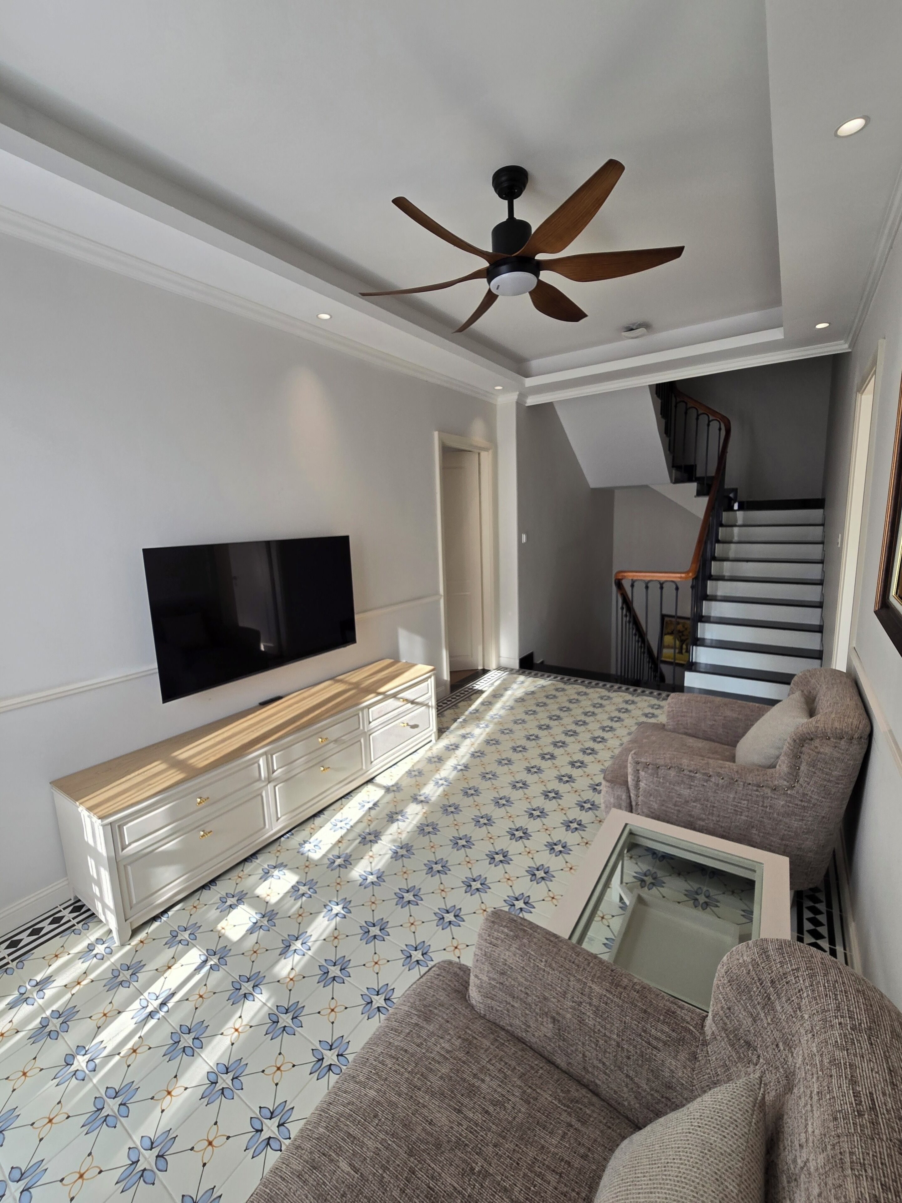 A living room with a flat-screen TV, a ceiling fan, a staircase, and patterned flooring.