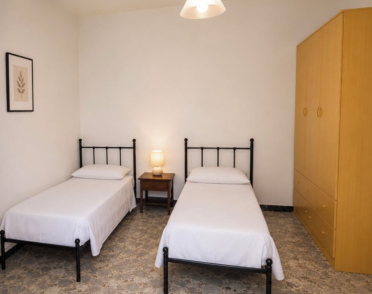 Et værelse med to enkeltsenge, et sengebord med lampe, et træskab og et billede i ramme på væggen.
