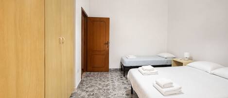 A hotel room with a wardrobe, a bed with a white bedspread, a bedside table with a lamp, and a tiled floor.