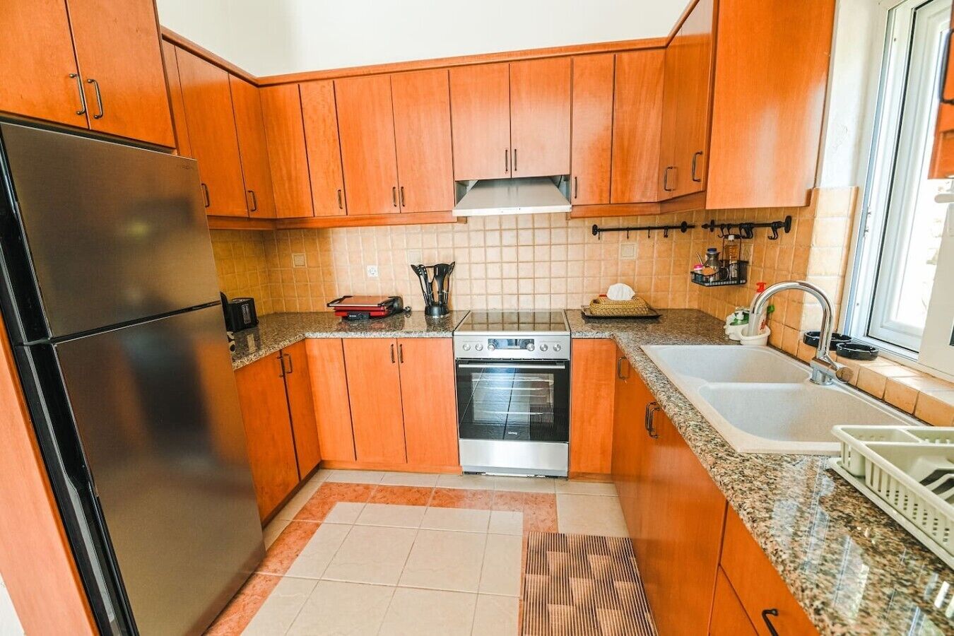 A compact kitchen with wooden cabinets, a stainless steel refrigerator, an oven, a sink, and a window providing natural light.