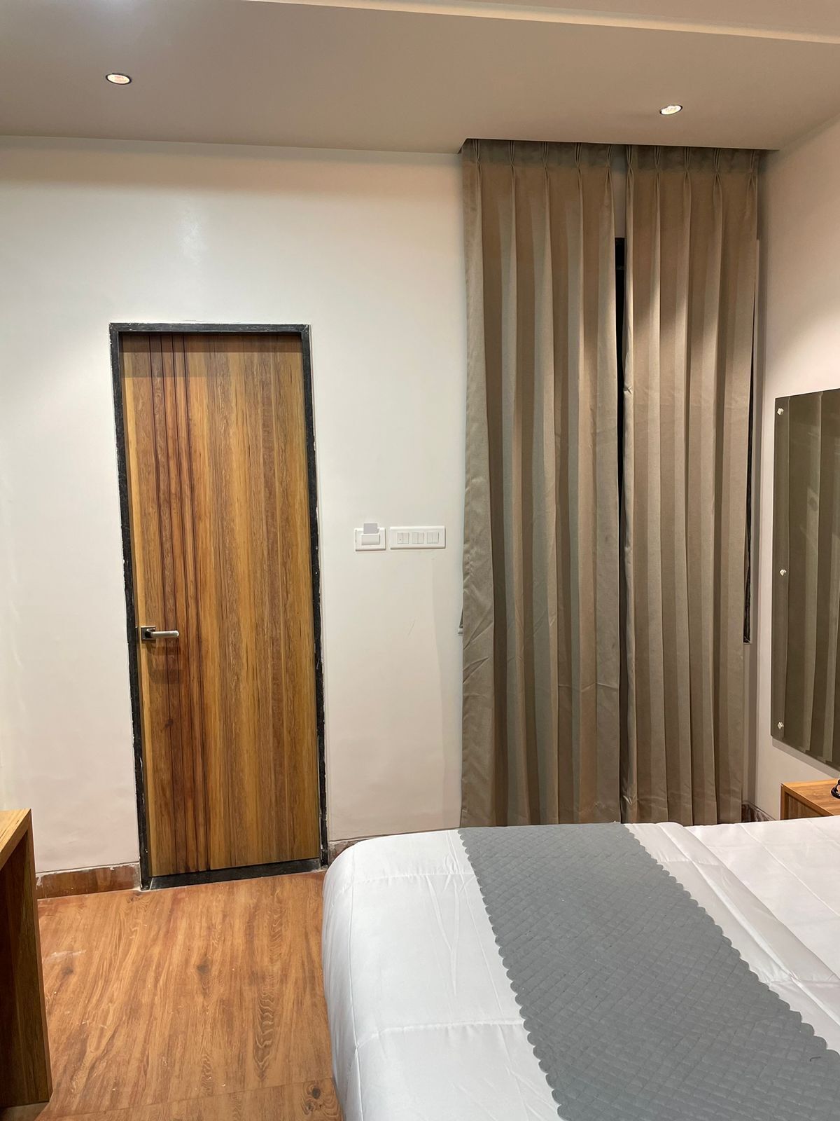 A bedroom with a wooden door, a bed with a grey and white bedspread, a mirror on the wall, and a window with curtains.
