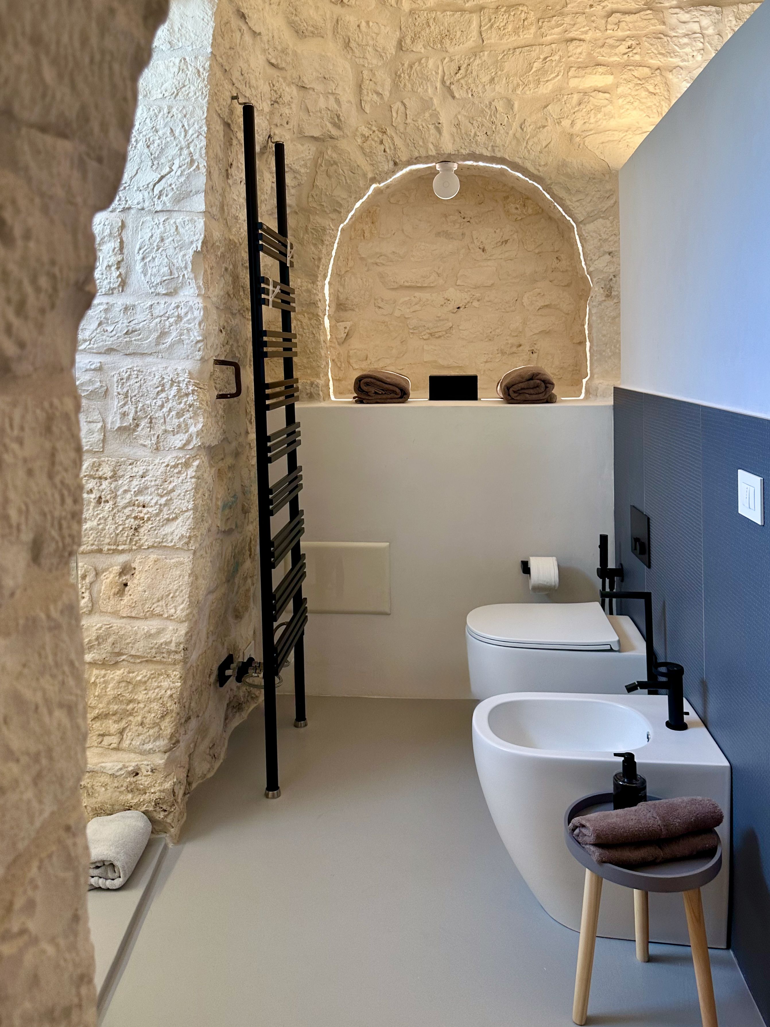 Un bagno moderno con parete in pietra, porta asciugamani nero, lavandino bianco e water.