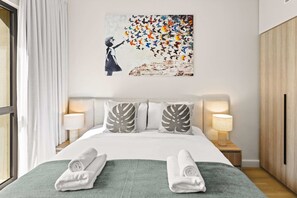 Ein Hotelzimmer mit einem Bett, zwei Nachttischlampen, einem Gemälde und zwei aufgerollten Handtüchern auf einer grünen Bettdecke.