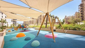 A children's play area with soft play surfaces, swings, and a slide, surrounded by buildings. - StayShort | Stylish 1BR Apartment | Near Burj Al Arab | Family-Friendly | Sleeps 4 (Dubai)