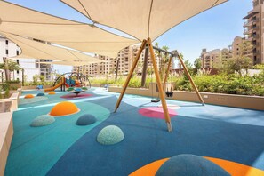 Ein Kinderspielbereich mit weichen Spielflächen, Schaukeln und einer Rutsche, umgeben von Gebäuden.