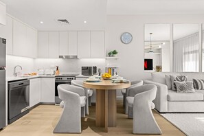 A modern kitchen with white cabinetry, a dining area with a round wooden table, and a living room with a grey sofa and decorative pillows. - StayShort | Stylish 1BR Apartment | Near Burj Al Arab | Family-Friendly | Sleeps 4 (Dubai)