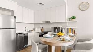 A modern kitchen with white cabinets, a stainless steel refrigerator, a dishwasher, a stove, and a microwave. There is a wooden dining table with chairs, a bowl of fruit, and a clock on the wall. - StayShort | Stylish 1BR Apartment | Near Burj Al Arab | Family-Friendly | Sleeps 4 (Dubai)