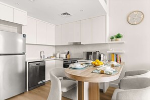 Eine moderne Küche mit weißen Schränken, einem Edelstahl-Kühlschrank, einer Spülmaschine, einem Herd und einer Mikrowelle. Ein Holztisch mit Stühlen, eine Schüssel mit Obst und eine Uhr an der Wand sind ebenfalls vorhanden.