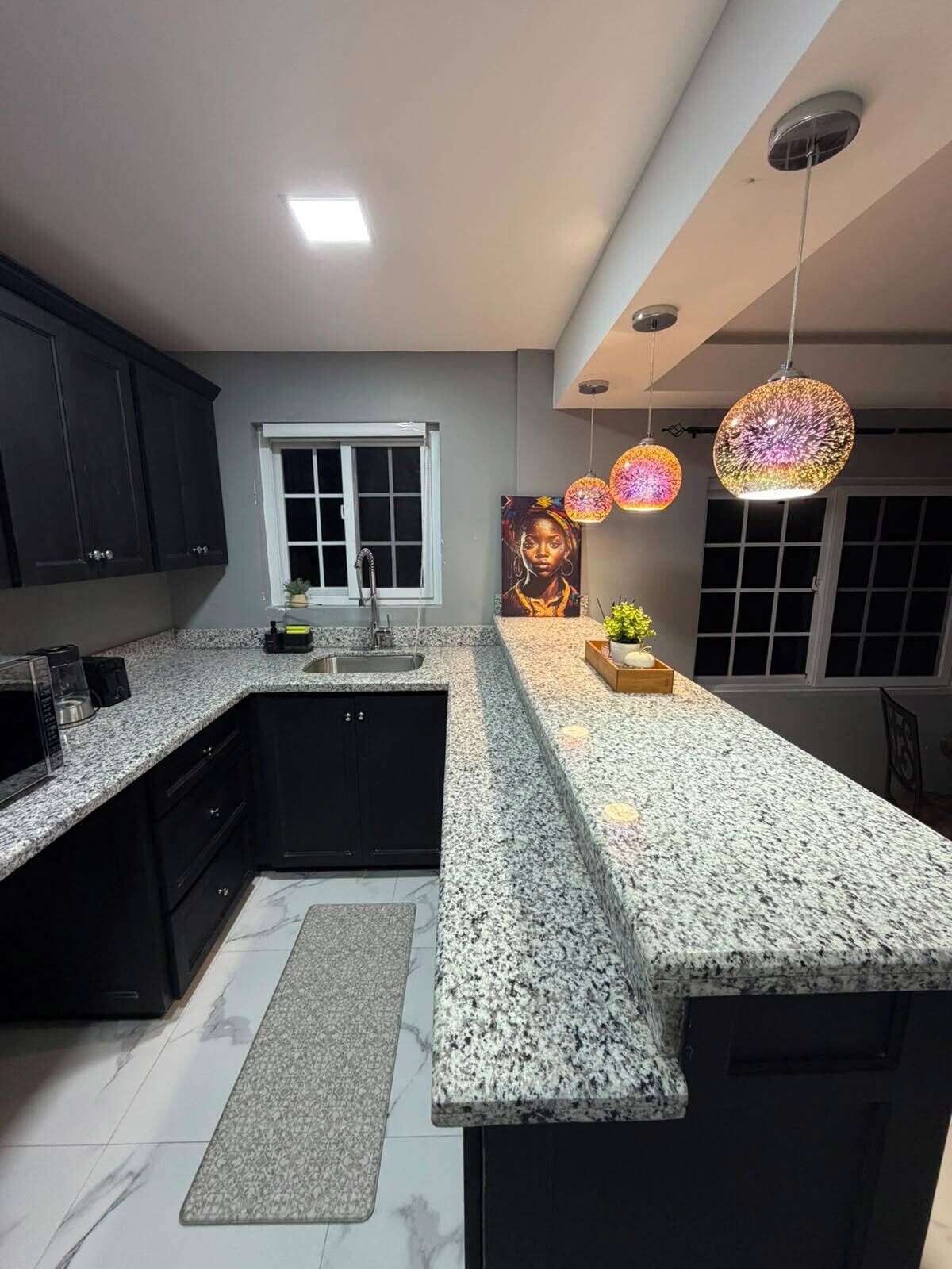 A kitchen with granite countertops, dark cabinets, a window, a sink, a microwave, and decorative hanging lights.