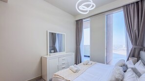 A modern hotel room with a large bed, a bedside table, a mirror, and a view of the city through the window. - Harbour Gate Residences - Creek Harbour (Dubai)