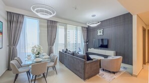 En moderne stue med sofa, spisebord og stole, et fladskærms-tv på væggen og et stort vindue med gardiner.