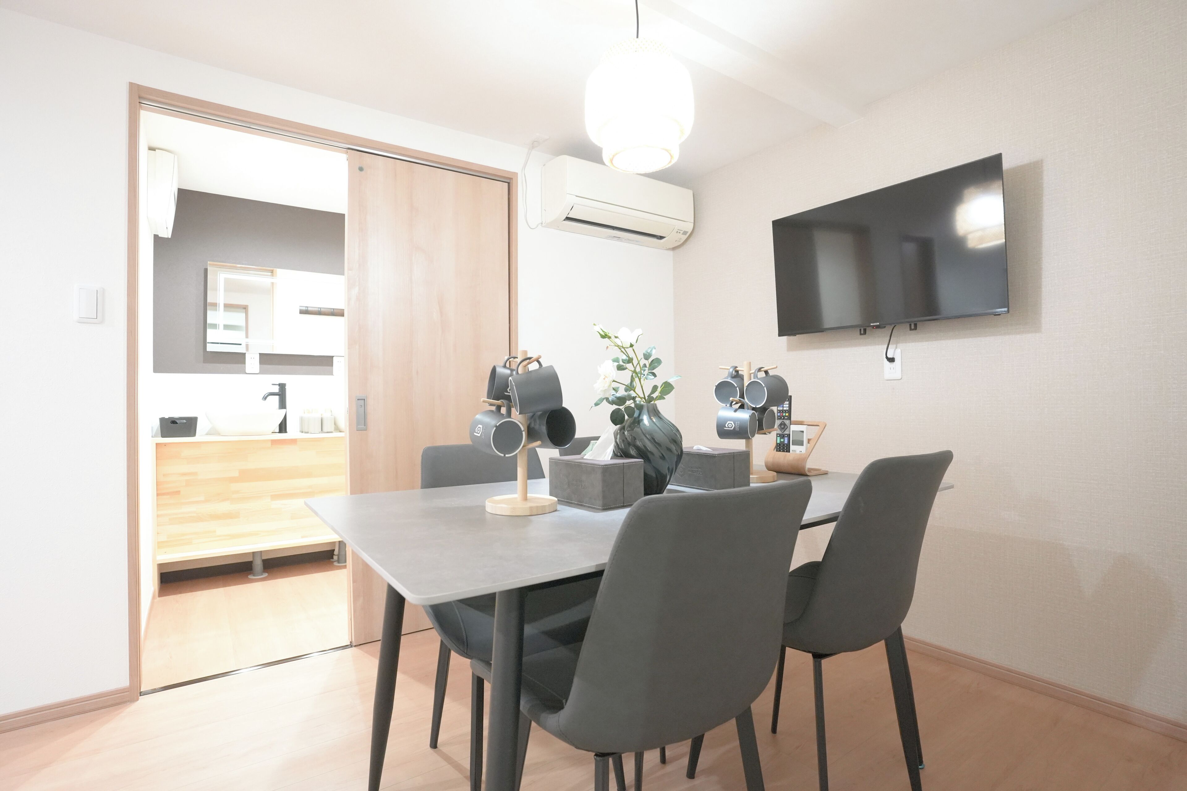 A modern dining area with a flat-screen TV mounted on the wall, a table with chairs, and a kitchen visible through an open door.