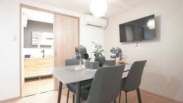 A modern dining area with a flat-screen TV mounted on the wall, a table with chairs, and a kitchen visible through an open door.