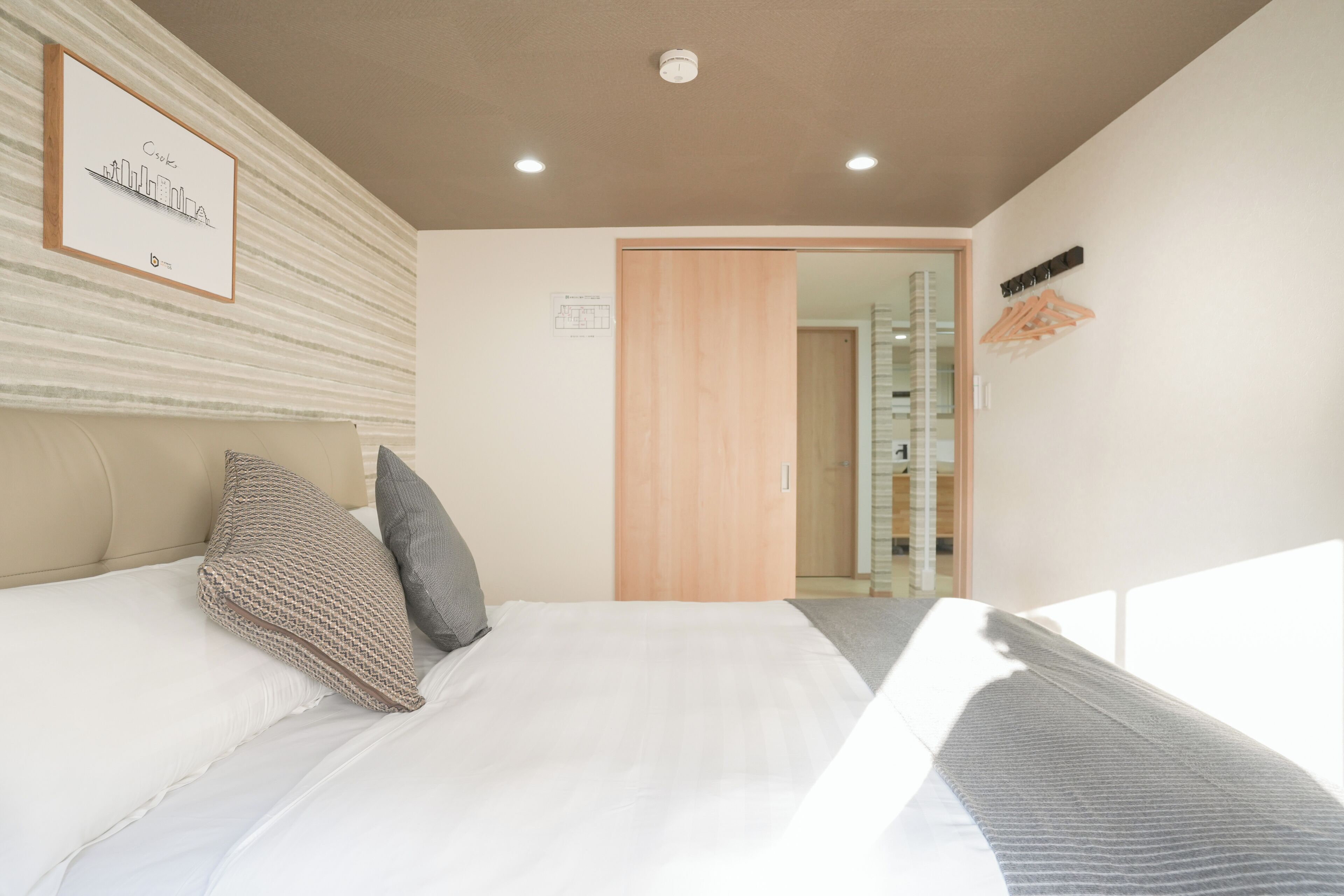 A hotel room with a bed, pillows, a framed picture on the wall, a wooden door, and a coat rack.