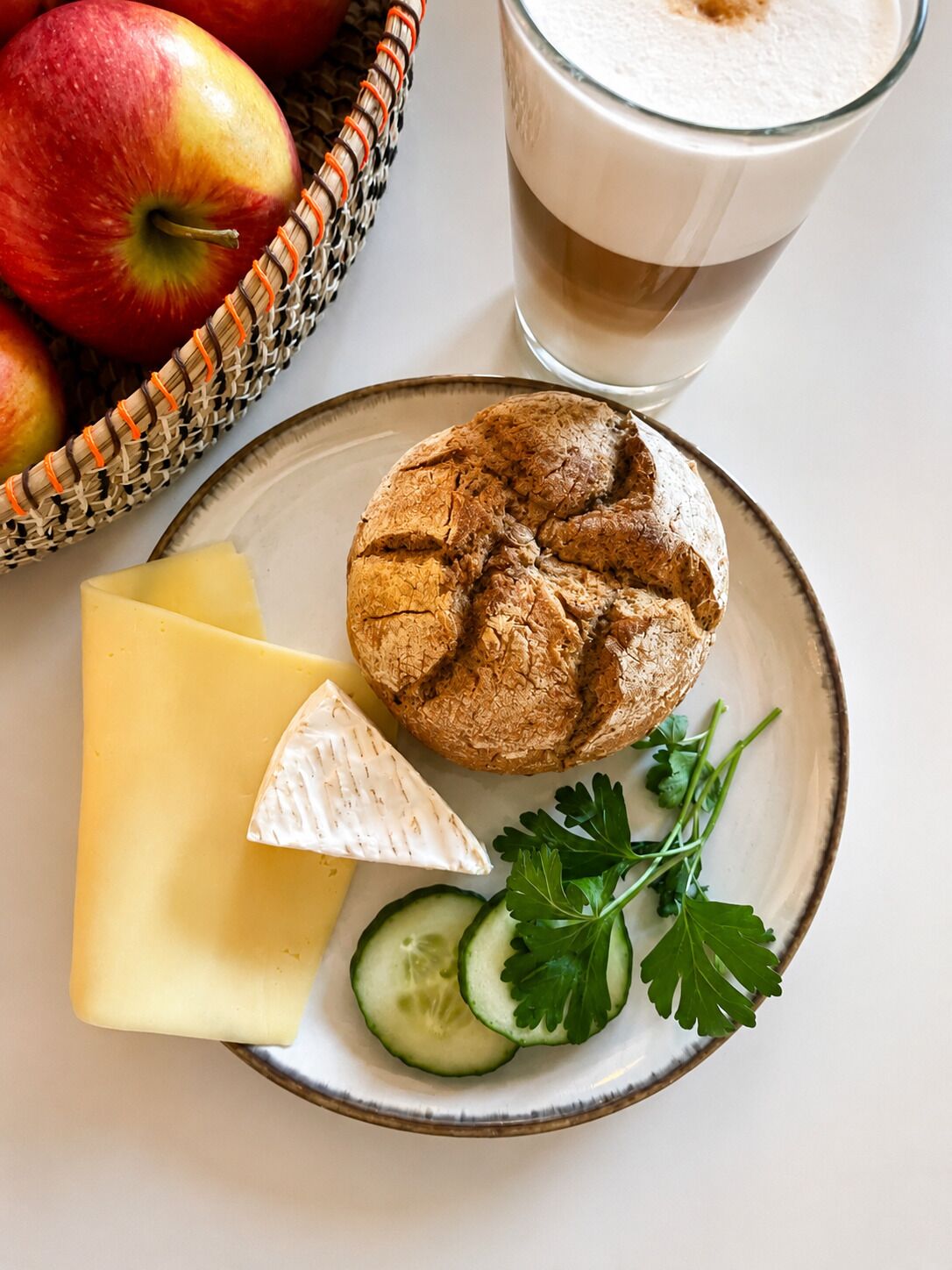 Un plat amb un muffin, formatge i rodanxes de pepino, un got de llet i una cistella de pomes.