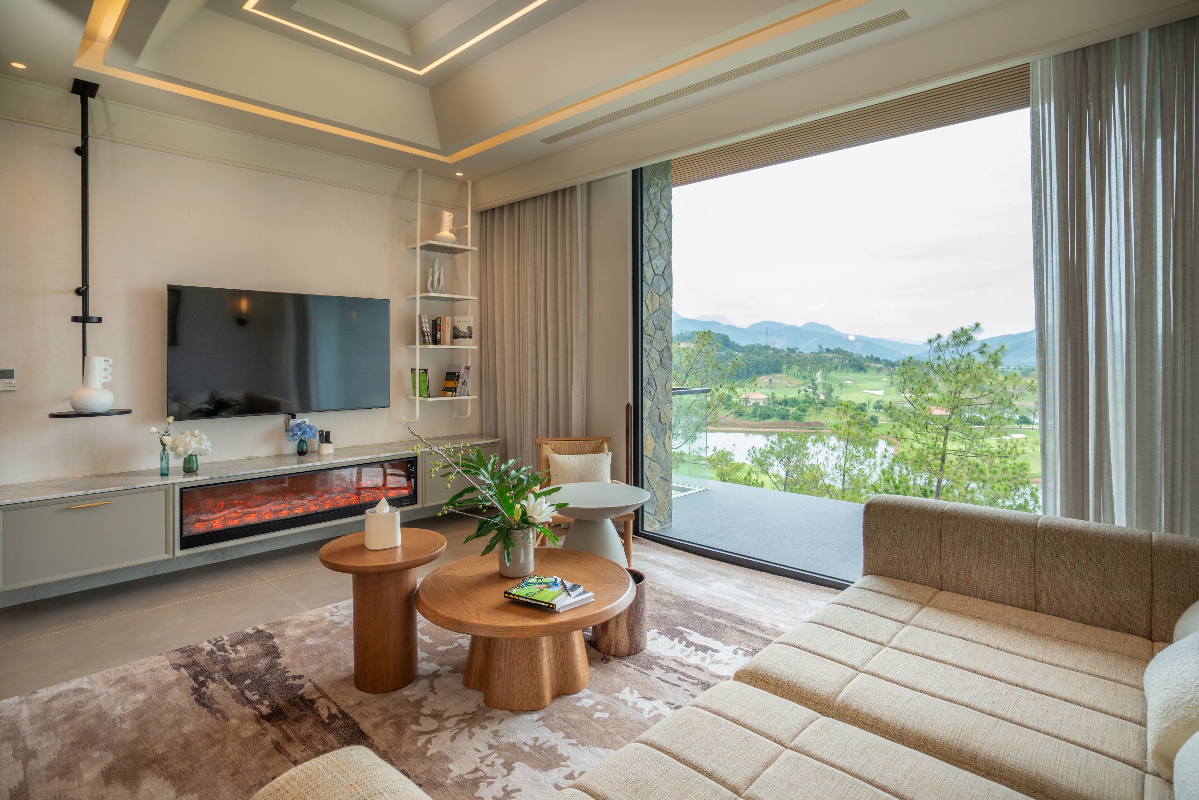 A modern living room with a flat-screen TV, a fireplace, a sofa, and a view of a green landscape.