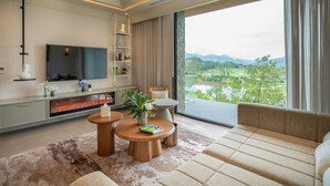 A modern living room with a flat-screen TV, a fireplace, a sofa, and a view of a green landscape.