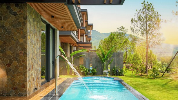 A swimming pool with a waterfall feature, surrounded by a well-maintained garden and a view of distant hills.