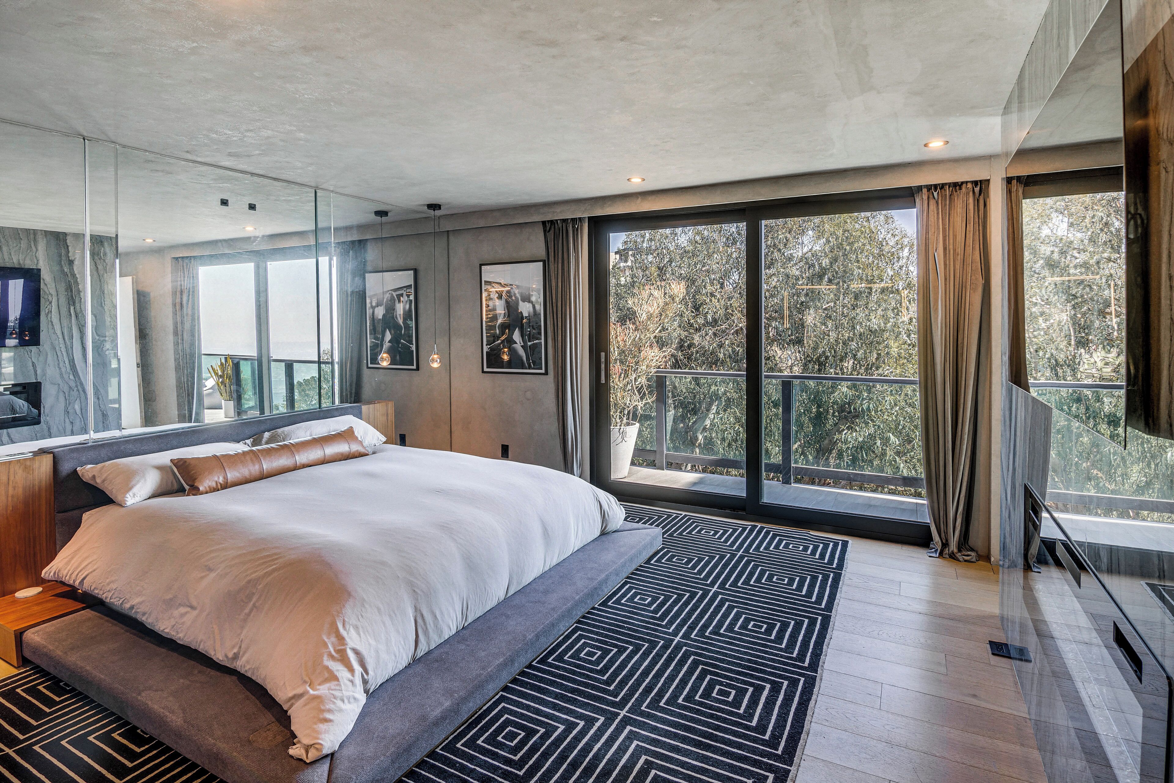 Ein modernes Schlafzimmer mit einem großen Bett, einer Glastrennwand und Blick auf Bäume durch große Fenster.