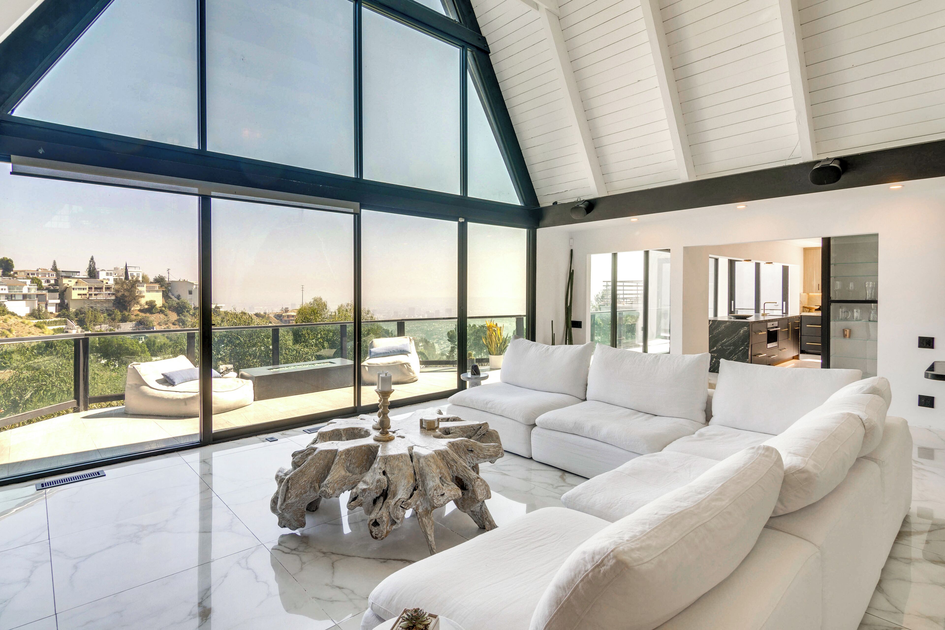 Un salón amplio con un sofá blanco grande, una mesa de centro de madera original y vistas a un paisaje urbano a través de ventanales amplios.