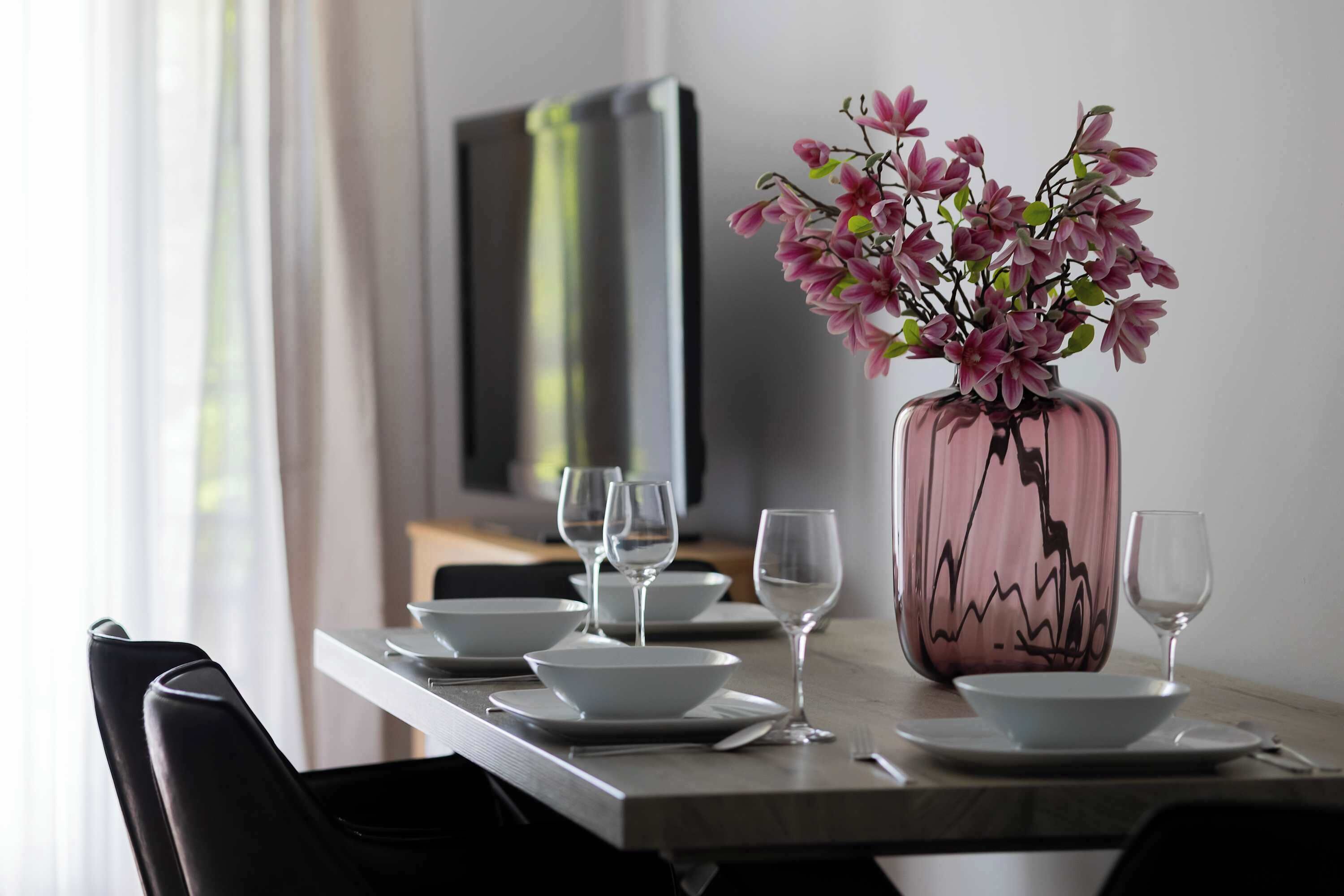 A dining table set with white plates, bowls, and wine glasses, with a vase of pink flowers in the center.