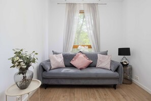 A modern living room with a grey sofa, a small side table, a floor lamp, and a window with sheer curtains. - Comfortable Apartment with a Balcony  Near the Airport (Warsaw)