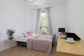 A bedroom with a bed, a desk with a laptop, a vase with flowers, and a window with curtains. - Comfortable Apartment with a Balcony  Near the Airport (Warsaw)