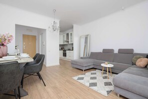 A modern living room with a grey sectional sofa, a dining area with a table and chairs, and a kitchen in the background. - Comfortable Apartment with a Balcony  Near the Airport (Warsaw)