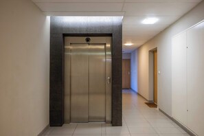 A hallway with a central elevator, white walls, and a tiled floor. - Comfortable Apartment with a Balcony  Near the Airport (Warsaw)