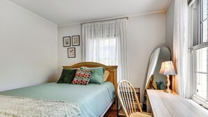 Quarto com cama, uma cadeira, escrivaninha e janela com cortinas.