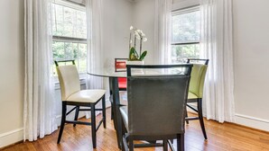 Uma sala de jantar com mesa de vidro rodeada por quatro cadeiras, duas janelas com cortinas brancas e uma orquídea em vaso sobre a mesa.