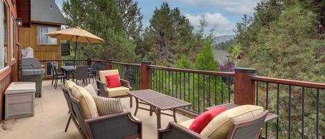 Une terrasse extérieure couverte, aménagée avec des meubles en osier, un barbecue et offrant une vue sur les arbres et les montagnes.