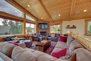 A spacious living room with a stone fireplace, leather sofas, and large windows offering a view of the outdoors. - 7 Min to Slopes! 3BD 2.5BA 5 Beds + Sofa Bed Cabin (Big Bear)