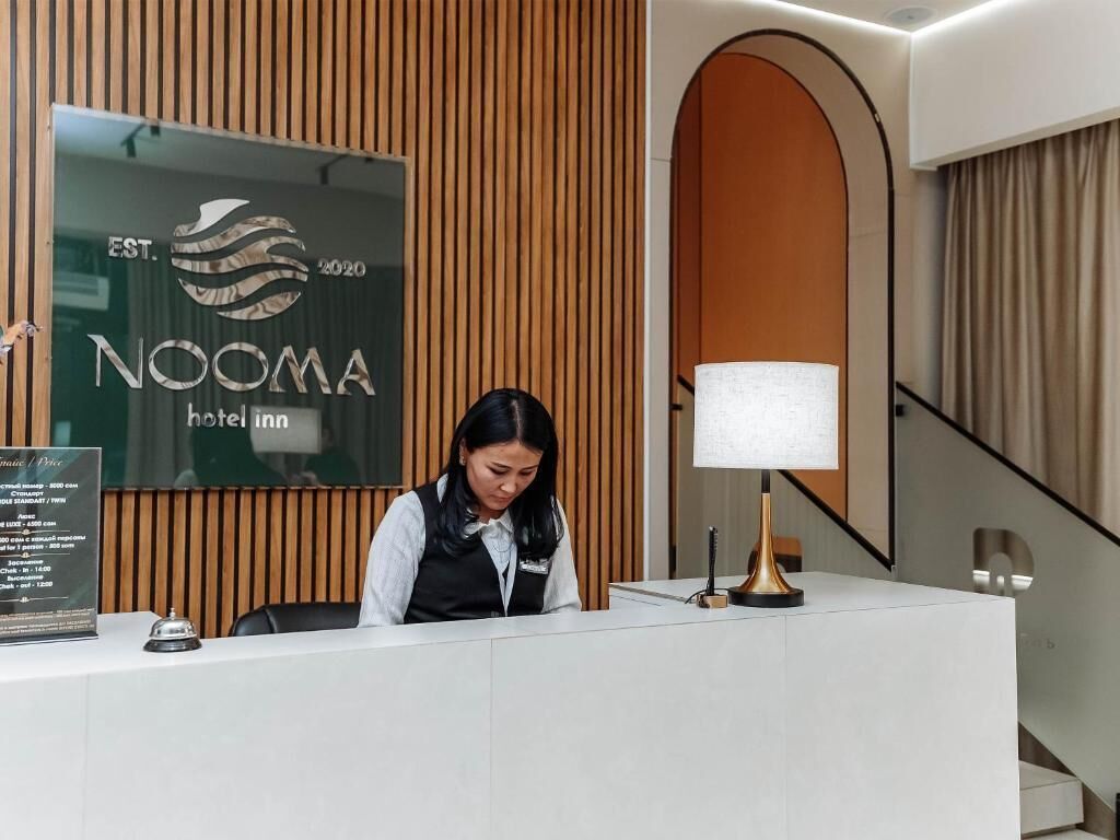 Un mostrador de recepción con el letrero del hotel NOOMA, una zona de recepción con una lámpara y una escalera al fondo.