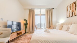 A hotel room with a bed, a TV, a desk, a chair, and a balcony with curtains. - HAVN - Studio in JVC with Pool View (Dubai)
