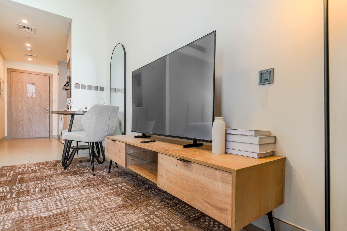 Un salon moderne avec un grand téléviseur à écran plat posé sur une console en bois, un fauteuil gris, une petite table avec un verre à vin et un tapis à motifs.