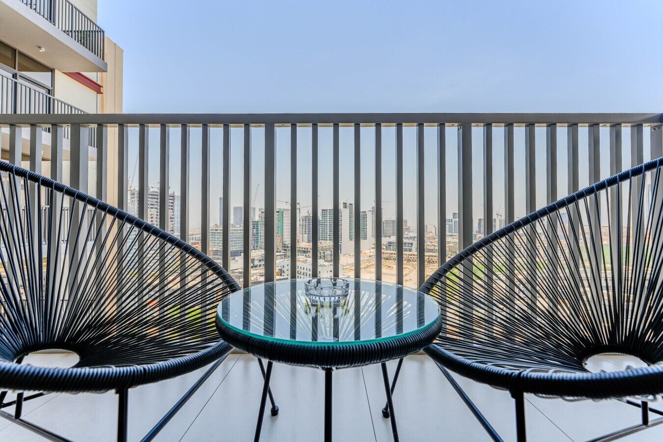 A balcony with a glass table and two chairs, overlooking a cityscape with buildings and construction.