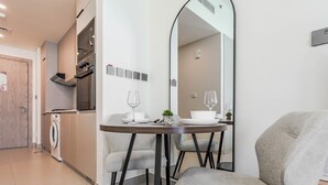 A modern kitchen with a built-in oven and microwave, a dining area with a round table and two chairs, and a large mirror reflecting the room. - HAVN - Studio in JVC with Pool View (Dubai)