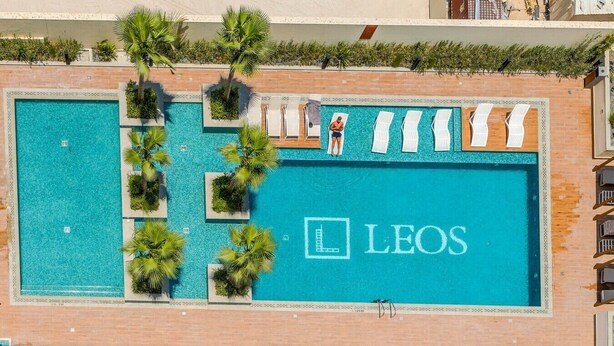 A swimming pool with the 'LEOS' logo, surrounded by palm trees and lounge chairs. - HAVN - Studio in JVC with Pool View (Dubai)
