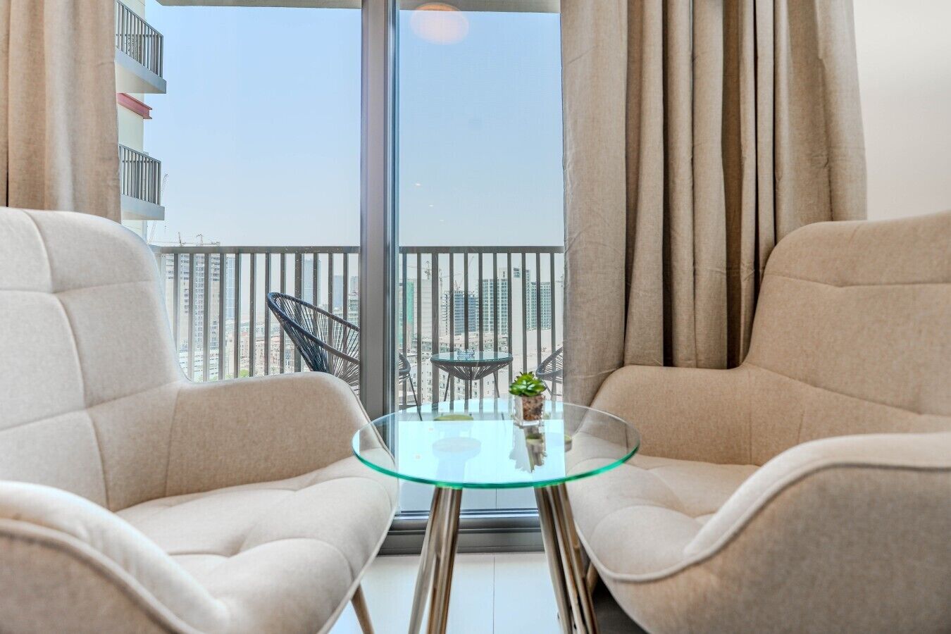 A balcony with two beige armchairs and a glass table, overlooking a cityscape.