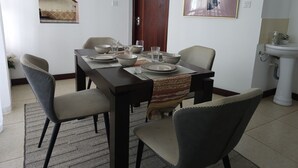 Comedor con mesa rectangular dispuesta para cuatro personas, sillas alrededor, un cuadro en la parte superior y una puerta a la izquierda.