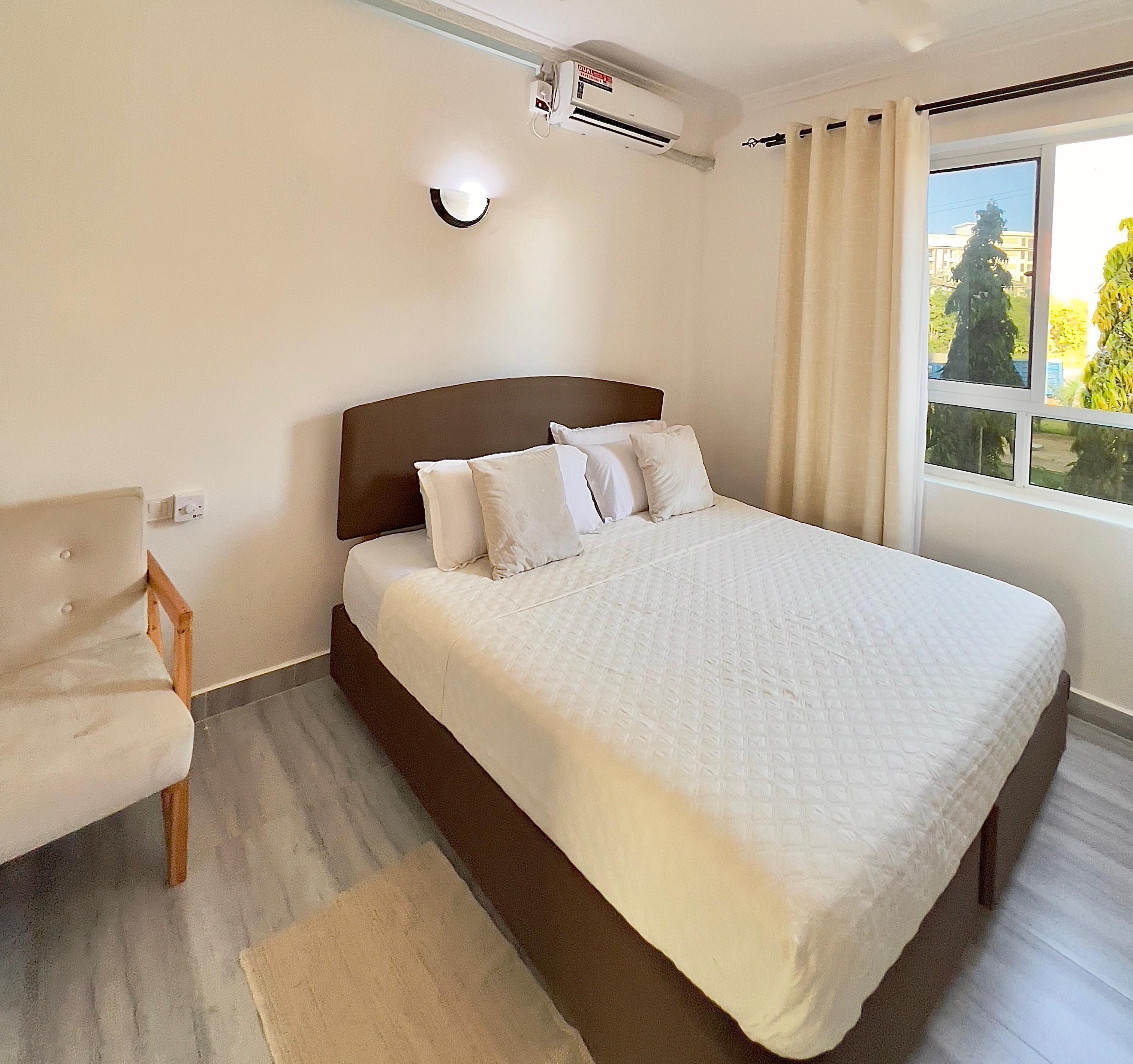 A hotel room with a bed, a chair, a window with curtains, and an air conditioning unit.