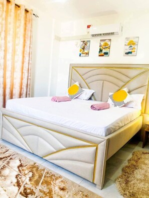 Una cama bien hecha con cabecera, aire acondicionado de pared y cuadros colgados encima.