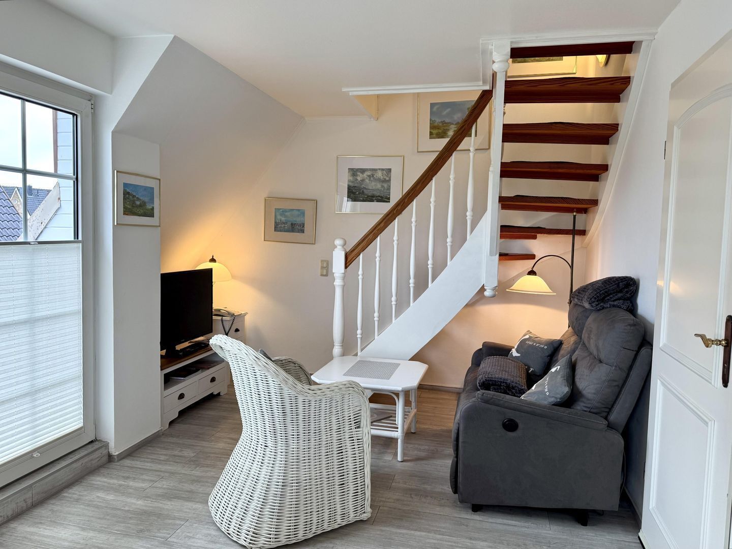A modern living room with a staircase, a sofa, a wicker chair, a TV, and framed pictures on the wall.