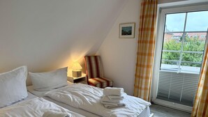 A hotel room with a bed, a chair, a nightstand, a lamp, a picture on the wall, and a window with curtains. - Kampstraße, Apartment 4a - Cozy 3-room vacation apartment with 2 balconies in a quiet (Sylt)