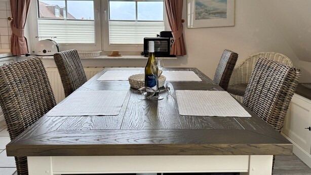 A dining area with a wooden table, wicker chairs, a bottle of wine, and a view of the outside through large windows. - Kampstraße, Apartment 4a - Cozy 3-room vacation apartment with 2 balconies in a quiet (Sylt)