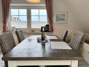 A dining area with a wooden table, wicker chairs, a bottle of wine, and a view of the outside through large windows. - Kampstraße, Apartment 4a - Cozy 3-room vacation apartment with 2 balconies in a quiet (Sylt)