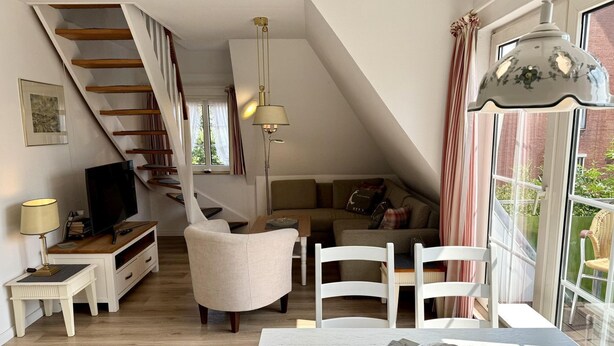 A modern living room with a staircase, a television, a dining area, and a large window. - Süderstraße, Apartment 12 – Landhaus - Cozy 3-room vacation rental for 4 (Sylt)