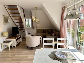 A modern living room with a staircase, a television, a dining area, and a large window. - Süderstraße, Apartment 12 – Landhaus - Cozy 3-room vacation rental for 4 (Sylt)